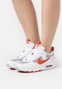 Vitaidens skor i vitt med röda accenter, i mesh och läder, rund tå och dämpade sulor. Utrustade med en distinkt logotyp och randiga dragöglor.