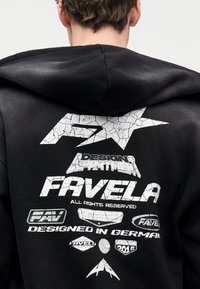 Άτομο που φοράει μαύρο φούτερ με κουκούλα με λευκά ραγισμένα γραφικά λογότυπα και κείμε νο, συμπεριλαμβανομένων των λέξεων "FAVELA", "DESIGN DEPARTMENT" και "DESIGNED IN GERMANY", στην πλάτη.
