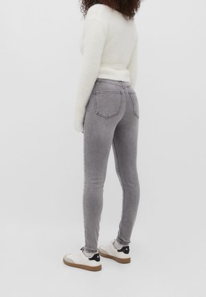 Mujer con jeans ajustados grises, blusa blanca de manga larga y peluda, y zapatillas blancas con detalles negros, de pie sobre un fondo blanco liso.