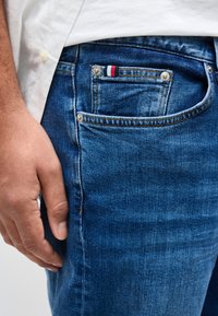 Κοντινό πλάνο του χεριού ενός ατόμου που ακουμπά δίπλα σε μπλε τζιν με μια μικρή κόκκινη, λευκή και μπλε ετικέτα στην τσέπη για κέρματα, φορώντας άσπρο πουκάμισο.