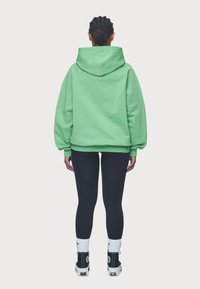 Sudadera verde de gran tamaño con capucha ajustable, combinada con legging negros y zapatillas altas, con calcetines blancos y acentos negros.