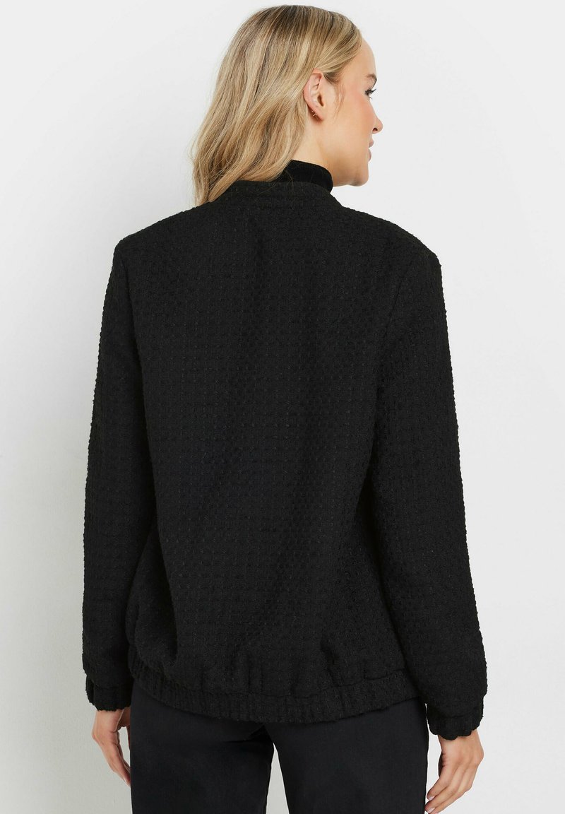 Svart texturerad jacka med hög krage, ribbade ärmslut och nederkant. Har ett subtilt våffelmönster och en avslappnad passform. Bakvy visas.