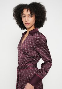 Φόρεμα πουκάμισο από σατέν σε μπορντό χρώμα με γιακά, ύφασμα με σχέδια, μακριά μανίκια και ζώνη που δένει στον ίδιο τόνο· γυαλιστερή υφή και μανσέτες σε αντίθεση.