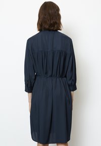 Vestito camicia blu navy a maniche lunghe. Tessuto morbido e fluido con parte posteriore arricciata e cintura in vita. Polsini con bottoni e leggera trasparenza all'orlo.