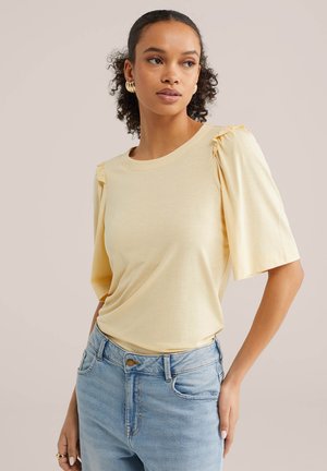 Donna che indossa una maglia giallo pallido con maniche a sbuffo e jeans a vita alta azzurri, con orecchini a cerchio dorati, che guarda di lato.