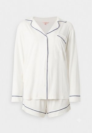 Femme debout pieds nus portant un haut de pyjama blanc à manches longues boutonné et un short assorti avec un passepoil foncé sur un fond uni.