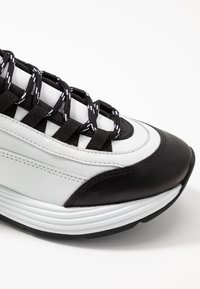 Sneaker bianco e nero con superiore in pelle texturizzata, spessa suola bianca e lacci neri a fantasia. Design della punta arrotondata.
