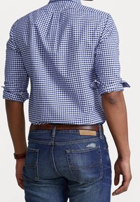 Camisa a cuadros azul y blanca con mangas remangadas, combinada con jeans de mezclilla azul oscuro, con un cinturón de cuero marrón y costuras visibles en los bolsillos.