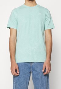 Mátově zelené bavlněné tričko s krátkým rukávem, kulatým výstřihem a malým bílým logem na hrudi. Měkká textura a uvolněný střih.