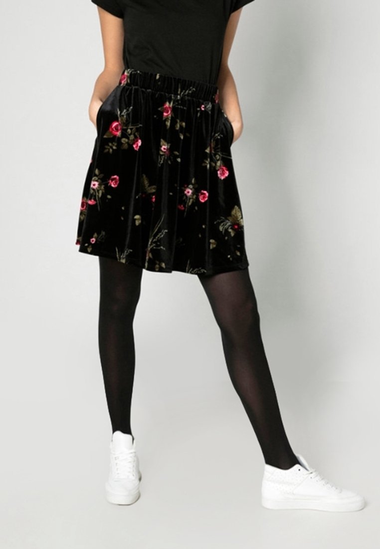 Femme portant une jupe en velours noire à fleurs, des collants noirs, des baskets blanches et un haut noir à manches courtes, debout les mains dans les poches.