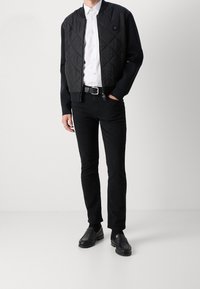 Blouson aviateur noir matelassé, chemise blanche à boutons, pantalon noir slim, ceinture noire et mocassins noirs. Tissu texturé avec un détail logo.