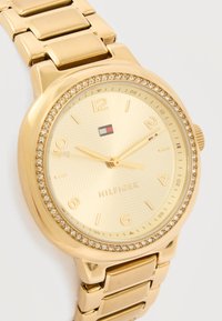 Χρυσό ρολόι χειρός Tommy Hilfiger με στεφάνη διακοσμημένη με κρύσταλλα, χρυσό βραχιόλι και χρυσό καντράν που δείχνει ώρα 10:09:10.