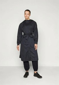 Vrouw in een zwarte, met een riem bevestigde trenchcoat met capuchon, draagt zwarte broek en zwarte sneakers, staat tegen een effen lichte achtergrond.