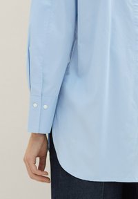 Chemise bleu clair à manches longues et boutons. Présente un ourlet arrondi et deux boutons blancs sur le poignet. Texture de tissu tissé lisse.