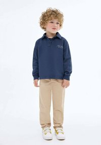 Námořnicky modrý polo s dlouhým rukávem a límečkem, béžové kalhoty a bílé tenisky s žlutými akcenty. Tkanina vypadá měkká a hladká.