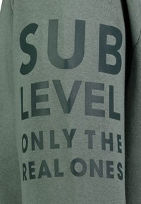 Groene sweatshirt met grote zwarte tekst die zegt "SUB LEVEL ONLY THE REAL ONES." De stof is zacht en heeft een lichte textuur.