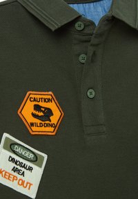 Polo de color verde oscuro con una tela texturizada. Presenta un parche hexagonal naranja etiquetado "CUIDADO WILD-DINO" y un parche blanco "ÁREA DE PELIGRO DINOSAURIO".