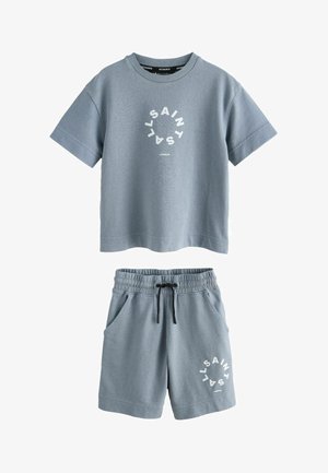 Dospievajúci chlapec stojí oblečený v modrom tričku a šortkách s okrúhlymi logami "SAINTS LONDON", bielymi ponožkami a bielymi teniskami.