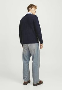 Navy blue cable-knit sweater with a rounded neckline and ribbed cuffs, paired with light blue jeans and brown boots, viewed from behind.