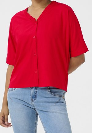 Frau trägt eine leuchtend rote Bluse mit Knöpfen und Rüschen am Ausschnitt sowie hellblaue Jeans, steht mit einem Arm hinter dem Rücken.