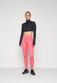 Top noir à manches longues coupé, associé à des leggings roses taille haute avec poches latérales, accompagné de chaussures de sport noires.