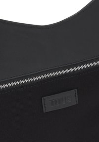 Sección de bolsa de tela negra con una cremallera horizontal plateada y una etiqueta negra cosida con relieve del nombre de la marca "TOUS".