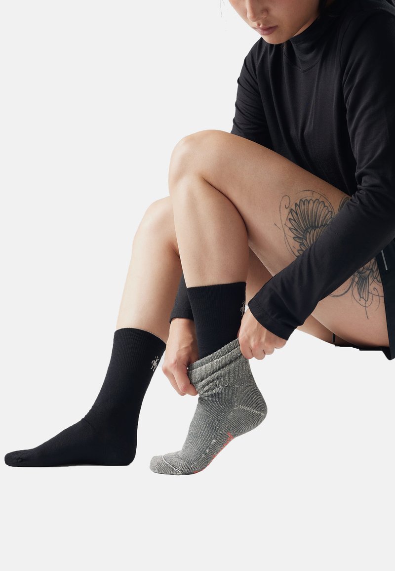 Persona che indossa una maglietta nera a maniche lunghe e pantaloncini, mettendo un calzino grigio sopra un calzino nero, con un grande tatuaggio floreale visibile sulla coscia.