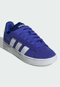 Le sneakers in suede viola presentano strisce bianche, lacci blu e una suola in gomma bianca. La linguetta ha un logo bianco su uno sfondo blu.