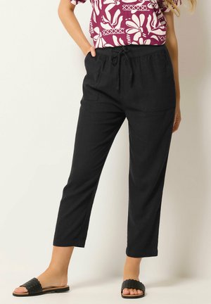 Kobieta nosząca czarne lniane spodnie z sznurkiem w pasie i przednimi kieszeniami, w połączeniu z czarnymi klapkami i wzorzystą bluzką z krótkim rękawem.