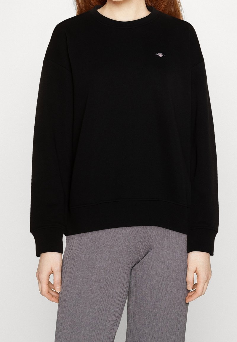 Femme portant un sweat-shirt noir uni à manches longues et un pantalon gris côtelé, debout devant un fond blanc avec les mains visibles.