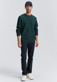 Pulover texturat de culoare verde închis, cu mâneci lungi și guler rotund; combinat cu pantaloni bleumarin și adidași albi, completat cu o șapcă închisă la culoare.
