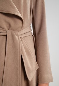 Beige kantavatka, jossa on teksturoitu kangas, leveä kaulus, pitkät hihat ja solmittava vyövyö keskellä.