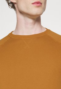 Sudadera de manga larga con cuello redondo en un cálido color mostaza, hecha de suave algodón, con costuras visibles y una textura lisa.