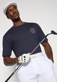 Camisa de manga curta azul-marinho com logótipo, cinto branco, calças brancas e luvas de golfe brancas, segurando um taco de golfe. Tecido suave, ajuste casual.