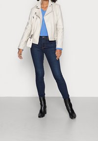 Kvinna iklädd en vit läderjacka, blå blus, mörka skinny jeans, svarta ankelboots och bär en vit axelväska.