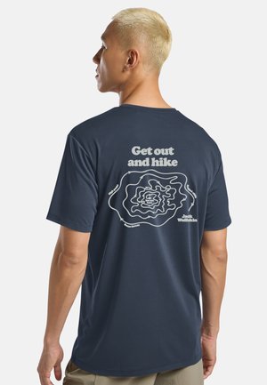 Mann mit kurzen blonden Haaren, der ein marineblaues T-Shirt mit weißer Konturenkarte und dem Text „Rausgehen und wandern“ auf dem Rücken trägt.