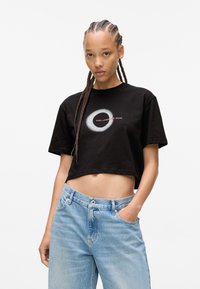 Melnā kokvilnas pusmugura T-kreklā ir apļa formas balts grafiks un teksts "KARL LAGERFELD JEANS." Apvienots ar gaiši ziliem džinsiem.