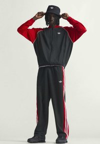 Svart och röd träningsoverall med vita ränder och logotyp, bestående av en jacka med dragkedja och byxor med resår i midjan. Levereras med en svart böjlig hatt.