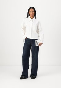 Camicia bianca con colletto e taschino sul petto, abbinata a jeans a gamba larga blu scuro. Gli accessori includono una pochette argentata e scarpe con tacco nere.