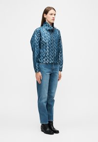 Γυναίκα που φοράει μπλε μπουφάν με φερμουάρ και σχέδια, μπλε τζιν και μαύρα μποτάκια, στέκεται μπροστά σε ένα απλό λευκό φόντο.