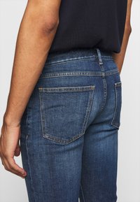 Jeans in denim blu scuro con un design aderente, dotati di due tasche posteriori, cuciture sottili e vita a media altezza. La texture appare liscia.