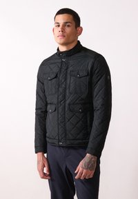 Black quilted jacket with a high collar, featuring two chest pockets, snap buttons, and textured fabric. Lightweight and fitted design.