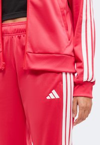 Άτομο που φοράει κόκκινη φόρμα Adidas με λευκές ρίγες, στέκεται με το δεξί χέρι χαλαρό στο πλευρό.
