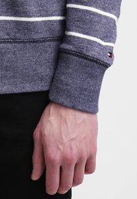 Rechterhand ontspannen aan de zijkant, gekleed in een blauw-wit gestreepte lange mouw met een klein rechthoekig logo vlakbij de pols.