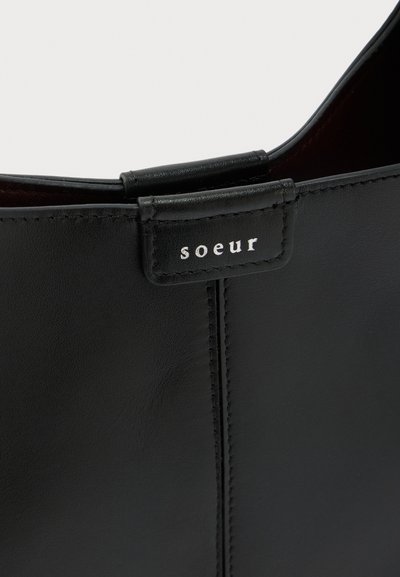 Gros plan sur une botte en cuir noir avec une couture cousue et un petit onglet portant le nom de la marque "soeur" en lettres blanches.