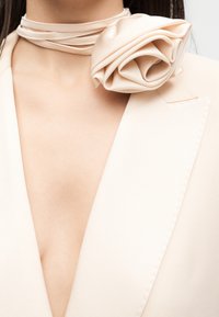 Μπεζ σακάκι με βαθύ V-λαιμό, σε συνδυασμό με ένα ασορτί σατέν τριανταφυλλένιο καρφίτσα και λεπτό περιδέραιο τσόκερ γύρω από το λαιμό μιας γυναίκας.