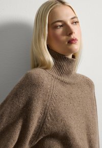 Maglione marrone lavorato a maglia con un alto colletto a coste, caratterizzato da una vestibilità ampia e una superficie testurizzata con sottili variazioni di colore.