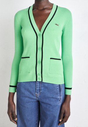 Slim-fit ribstrikket mintgrøn cardigan med sort kant, knapper foran, to forlommer, båret med blå jeans på en mørkhudet person.