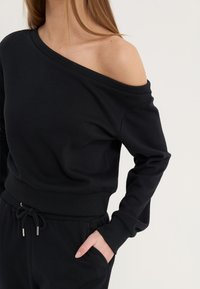 Sweat-shirt noir à épaules dénudées avec des manches longues, des poignets côtelés et une taille côtelée ; en coton doux avec une coupe décontractée.