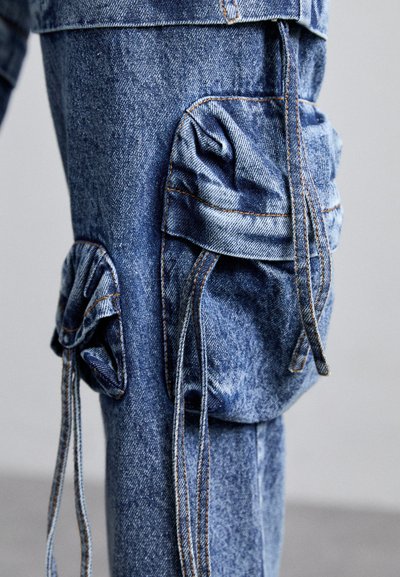 Pantaloni cargo din denim de culoare albastră deschisă, cu buzunare adunate, detalii cu șnur și cusături contrastante. Prezintă o croială relaxată și un material texturat.